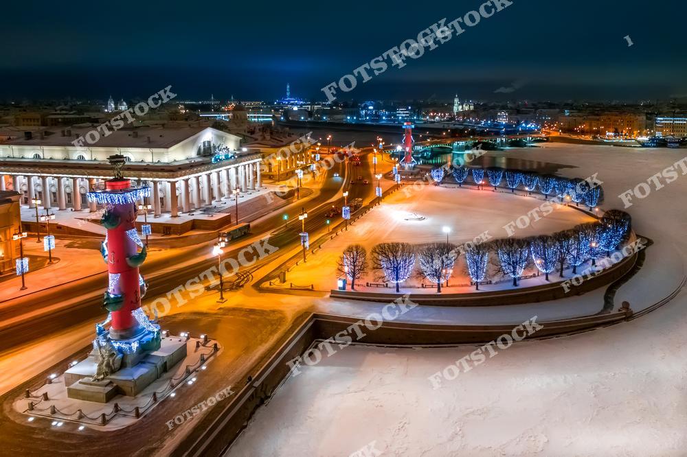 На фотографии изображена Стрелка Васильевского острова в Санкт-Петербурге, украшенная для праздников. Яркие огни гирлянд и освещенные ростральные колонны создают атмосферу зимнего волшебства. Заснеженная площадь и окрестности отражаются в ночных огнях города. Этот снимок с высоты передает красоту зимнего Санкт-Петербурга, его историческую атмосферу и праздничное убранство.